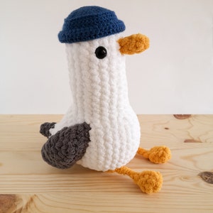 May include: A handmade white crochet seagull plush toy with a blue hat, gray wings, and yellow feet and beak. The seagull has a black eye and is sitting on a wooden surface.