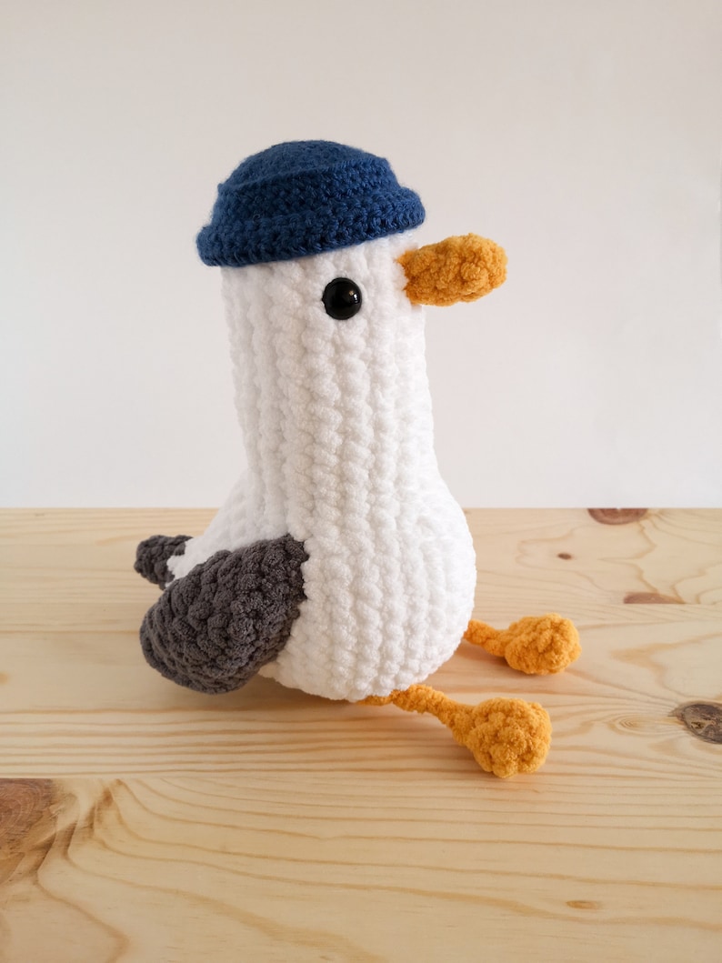 May include: A handmade white crochet seagull plush toy with a blue hat, gray wings, and yellow feet and beak. The seagull has a black eye and is sitting on a wooden surface.