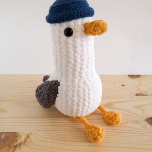 May include: A handmade white crochet seagull plush toy with a blue hat, gray wings, and orange feet and beak. The seagull has a round body and is sitting on a wooden surface. The plush toy has black eyes.