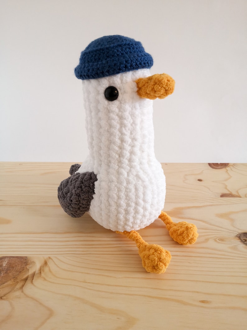 May include: A handmade white crochet seagull plush toy with a blue hat, gray wings, and orange feet and beak. The seagull has a round body and is sitting on a wooden surface. The plush toy has black eyes.