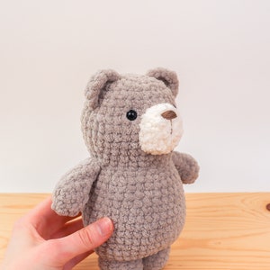 Może przedstawiać: Ręcznie robiony szary szydełkowy miś z białym pyskiem i brązowym nosem. Miś ma czarne oczy ochronne i małe uszy. Miś stoi na drewnianej powierzchni.