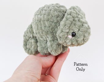 May include: A sage green crochet animal with black eyes and a pink nose. The soft toy is held in a hand. The image includes the text "Pattern Only".