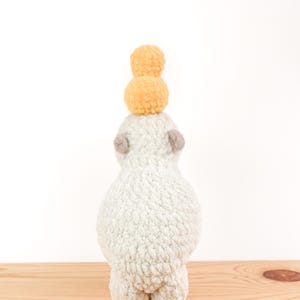 May include: A handmade, plush alpaca toy in a light gray color, with small gray ears and feet. Two yellow pom-poms are stacked on top of the alpaca's head. The toy is positioned on a wooden surface against a white background.