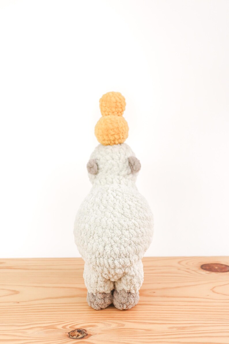 May include: A handmade, plush alpaca toy in a light gray color, with small gray ears and feet. Two yellow pom-poms are stacked on top of the alpaca's head. The toy is positioned on a wooden surface against a white background.