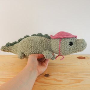 Op de afbeelding: Een groene gehaakte alligator knuffel met een roze gehaakte hoed. De alligator ligt op een houten oppervlak.