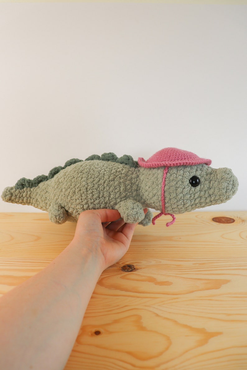 Op de afbeelding: Een groene gehaakte alligator knuffel met een roze gehaakte hoed. De alligator ligt op een houten oppervlak.