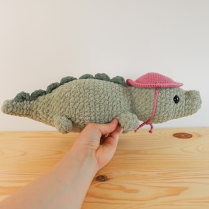 Op de afbeelding: Een gehaakt groen alligator knuffeldier met een roze gehaakte zonnehoed. De alligator heeft zwarte ogen en een klein roze strikje.