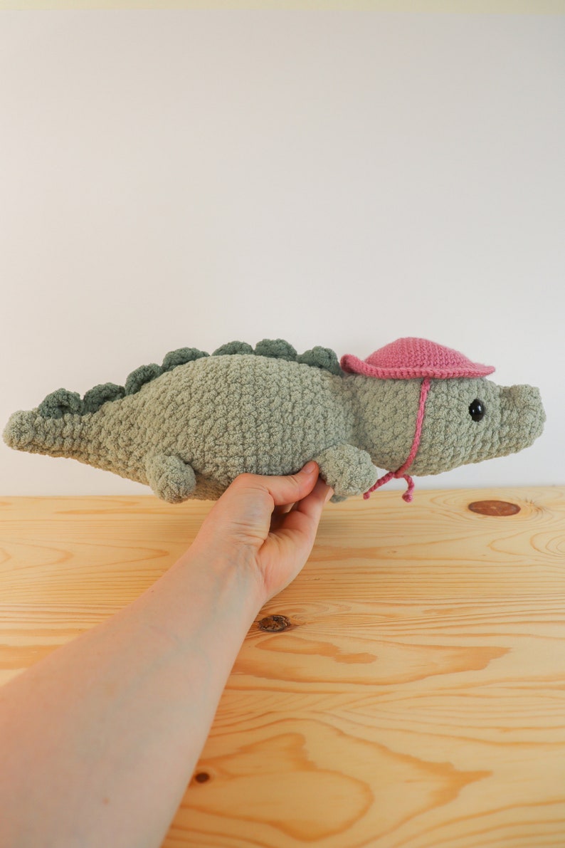 Op de afbeelding: Een gehaakt groen alligator knuffeldier met een roze gehaakte zonnehoed. De alligator heeft zwarte ogen en een klein roze strikje.