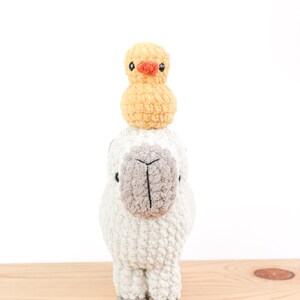May include: A handmade, crocheted llama with a small, yellow crocheted duck perched on its head. The llama is primarily off-white with a gray face and feet. The duck has an orange beak and black eyes. The toys are displayed on a light wood surface.