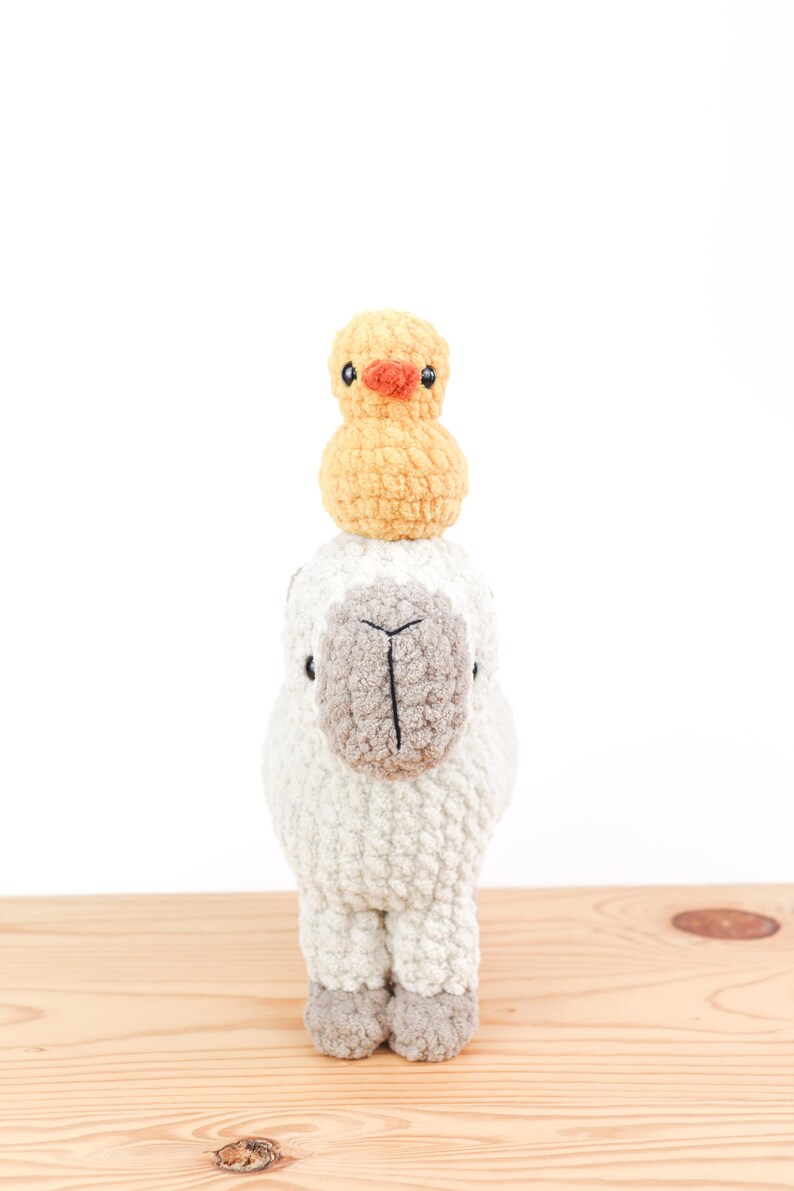 May include: A handmade, crocheted llama with a small, yellow crocheted duck perched on its head. The llama is primarily off-white with a gray face and feet. The duck has an orange beak and black eyes. The toys are displayed on a light wood surface.