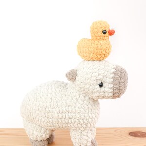 May include: A handmade crochet stuffed animal of a sheep with a small yellow duck perched on its head. The sheep is primarily white with gray accents on its head, ears, and feet. The duck has an orange beak.
