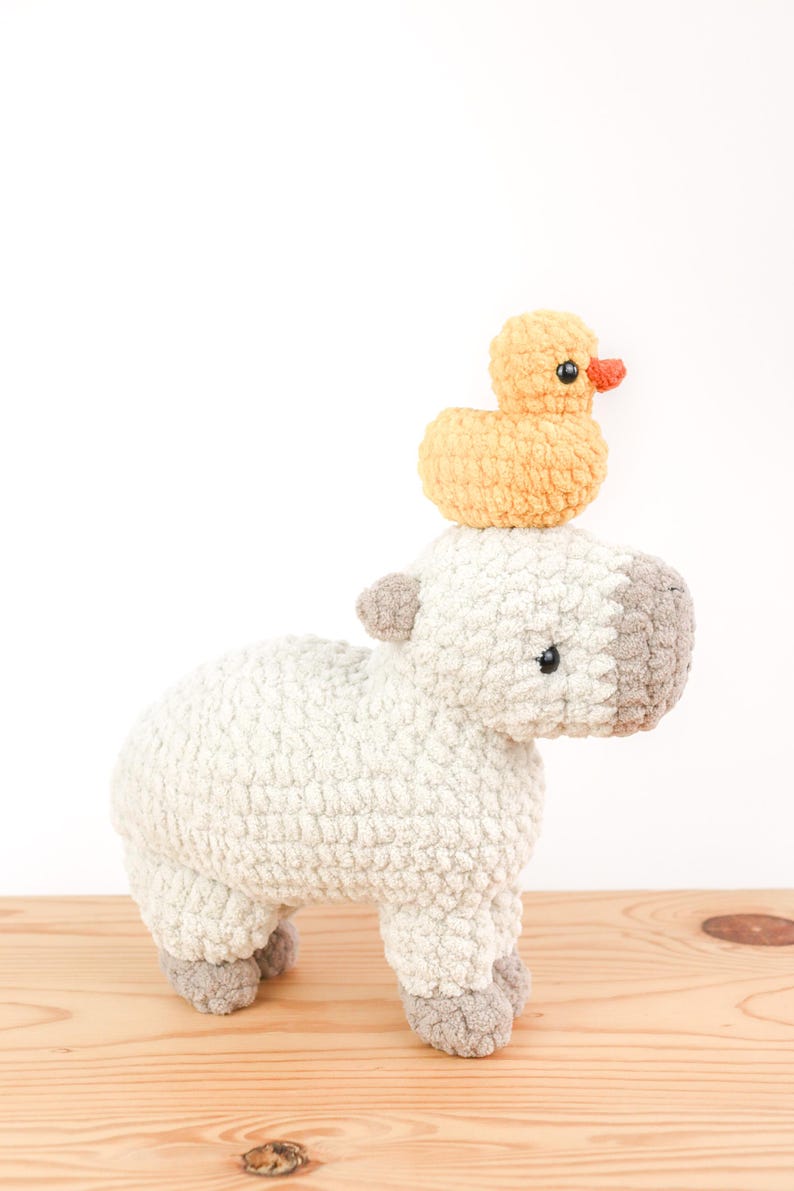 May include: A handmade crochet stuffed animal of a sheep with a small yellow duck perched on its head. The sheep is primarily white with gray accents on its head, ears, and feet. The duck has an orange beak.