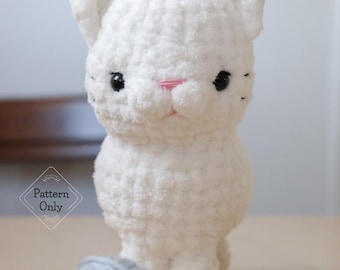 May include: A white crocheted cat toy with black eyes and a pink nose. The cat is sitting on a wooden surface next to a ball of grey yarn.