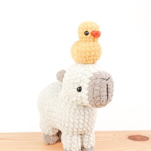 May include: A handmade, white and gray crochet llama with a small yellow duck perched on its head. The llama has black eyes and gray accents on its ears, nose, and feet. The duck has an orange beak. The toys are on a wooden surface.