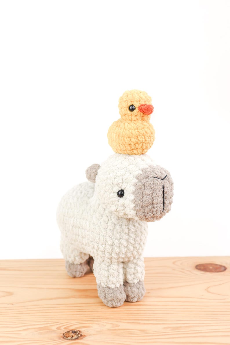 May include: A handmade, white and gray crochet llama with a small yellow duck perched on its head. The llama has black eyes and gray accents on its ears, nose, and feet. The duck has an orange beak. The toys are on a wooden surface.