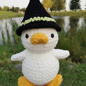 May include: White crocheted duck wearing a black witch hat with a green trim. The duck has yellow feet and a yellow beak.