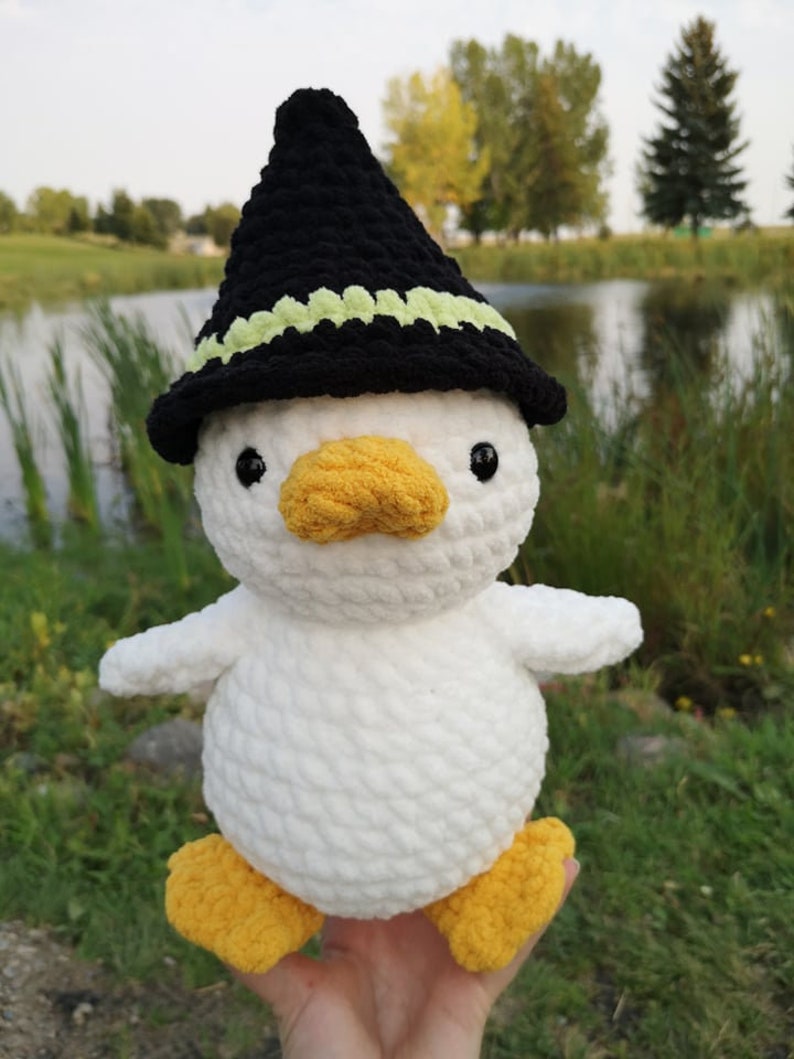 May include: White crocheted duck wearing a black witch hat with a green trim. The duck has yellow feet and a yellow beak.