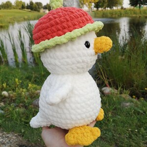 May include: White crocheted plush toy duck wearing a red and green crocheted apple hat. The duck has yellow feet and black eyes.