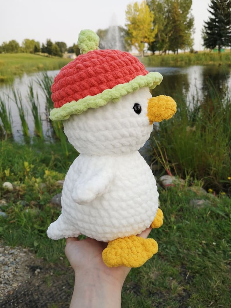 May include: White crocheted plush toy duck wearing a red and green crocheted apple hat. The duck has yellow feet and black eyes.
