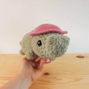 Op de afbeelding: Een gehaakt pluche speelgoed in de vorm van een neushoorn met een roze gehaakte hoed. Het speelgoed wordt vastgehouden door een hand.