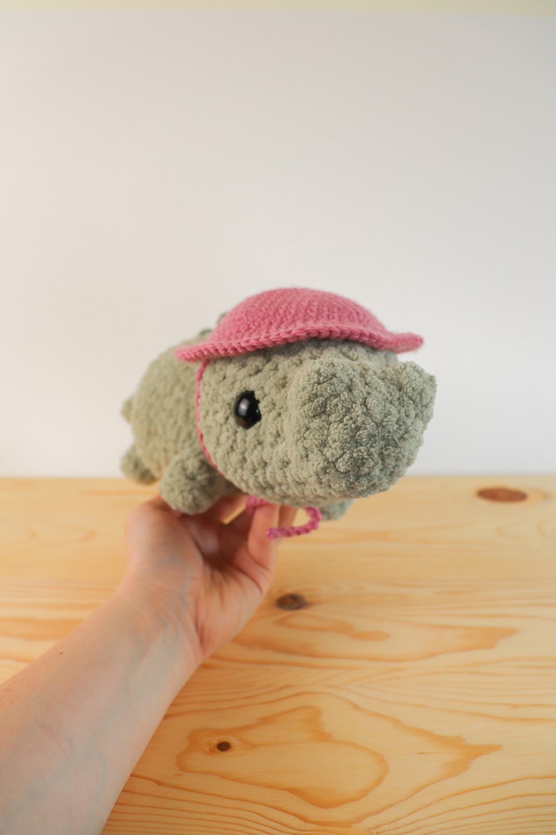 Op de afbeelding: Een gehaakt pluche speelgoed in de vorm van een neushoorn met een roze gehaakte hoed. Het speelgoed wordt vastgehouden door een hand.