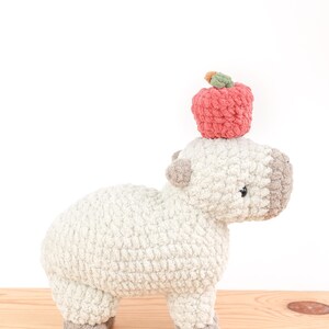 May include: A handmade, plush, light gray capybara with a light brown face and feet. A red crocheted apple with a green stem sits atop the capybara's head. The stuffed animal is on a wooden surface.