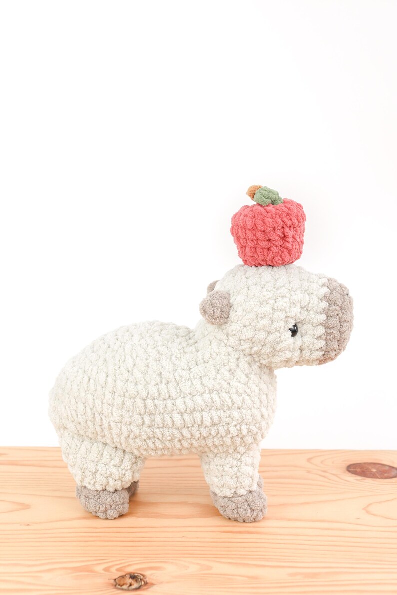 May include: A handmade, plush, light gray capybara with a light brown face and feet. A red crocheted apple with a green stem sits atop the capybara's head. The stuffed animal is on a wooden surface.