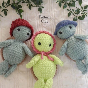May include: Three crocheted turtle amigurumi toys. One is green with a pink bonnet, one is blue with a blue hat, and one is gray with a red hat. The toys are sitting on a white surface.