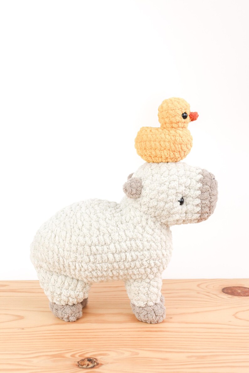 May include: A handmade crochet capybara plush toy in light gray with gray accents, topped with a small yellow crochet duck with an orange beak. The plush toys are on a wooden surface.