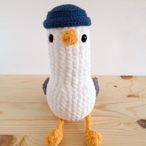 May include: A handmade, white crochet seagull plush toy with a blue hat, black eyes, and an orange beak and feet. The seagull has gray wings and is sitting on a wooden surface. This soft toy is perfect for children.