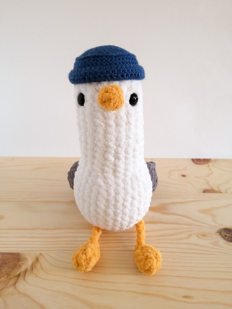 May include: A handmade, white crochet seagull plush toy with a blue hat, black eyes, and an orange beak and feet. The seagull has gray wings and is sitting on a wooden surface. This soft toy is perfect for children.