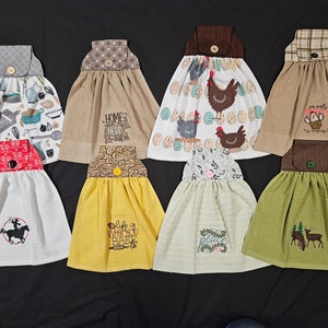 May include: Eight kitchen towels with different designs and colours. The towels are hanging against a black background. The designs include chickens, a cowboy on a horse, a deer, and a bear. The towels are all made from a soft, absorbent material.