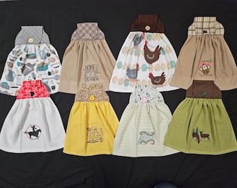 Op de afbeelding: Acht keukendoeken met verschillende ontwerpen en kleuren. De handdoeken hangen tegen een zwarte achtergrond. De ontwerpen omvatten kippen, een cowboy op een paard, een hert en een beer. De handdoeken zijn allemaal gemaakt van een zacht, absorberend materiaal.