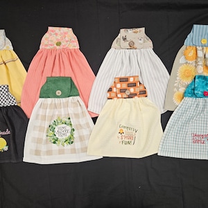 May include: Eight different kitchen towels with a pocket and button. The towels are in various colors and patterns, including yellow, pink, gray, beige, black, and blue. Each towel has a different embroidered design, including "Lemonade", "Home Sweet Home", "Campfire S'more Fun", and "Happiness is Kettle".
