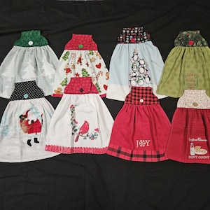 May include: Eight Christmas-themed dish towels with different designs. The towels are white with red, green, and blue accents. Some of the towels have embroidered designs, such as Santa Claus, a cardinal, and a snowman. Others have printed designs, such as a gingerbread house, a candy cane, and a Christmas tree. The towels are all trimmed with a red and white checkered border.