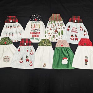 May include: A collection of Christmas-themed hanging kitchen towels. Each towel features a unique design, including gnomes, Santa, and festive patterns. The towels have decorative tops with plaid, polka dot, and striped fabrics.