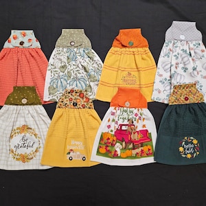 May include: Eight kitchen towels with autumn-themed designs. The towels are yellow, orange, green, and white with various patterns and text. The text includes "Be Grateful", "Happy Harvest", "Gather Together", "Hello Fall", and "Happy Harvest".