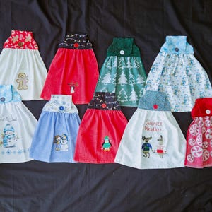 May include: A collection of decorative kitchen towels with festive holiday designs. The towels feature various patterns, including gingerbread men, snowmen, penguins, and candy cane prints. Each towel has a button closure and a patterned top.