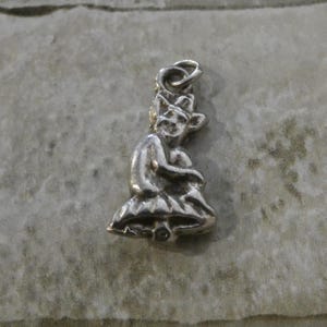 May include: A small, silver-toned charm in the shape of a seated goblin or imp. The figure has a pointed hat, a smiling face, and is holding its knees. The charm has a loop at the top for attaching to a necklace or bracelet.