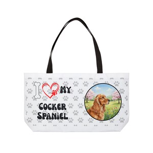 Może przedstawiać: Biała torba typu tote z czarnymi uchwytami. Zawiera ilustrację Cocker Spaniel w okrągłej ramce. Torba ma wzór łap i napis "I love my Cocker Spaniel" w kolorze czarnym i czerwonym.