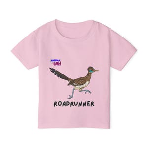 May include: A pale pink t-shirt with a cartoon roadrunner in brown, blue, and white. The word "ROADRUNNER" is printed in black below the image. The words "AMERICA WILD" are printed above the roadrunner.