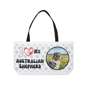 Może przedstawiać: Biała torba typu tote z czarnymi uchwytami z napisem "I ❤️ MY AUSTRALIAN SHEPHERD" i śladami łap. Po prawej stronie znajduje się okrągły obraz psa rasy Australian Shepherd na polu.