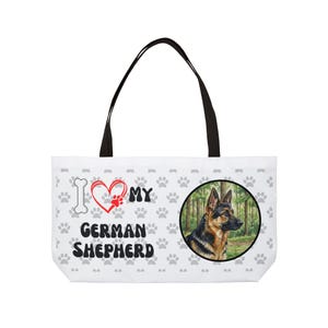 Może przedstawiać: Biała torba typu tote z czarnymi uchwytami. Zawiera ilustrację owczarka niemieckiego w leśnej scenerii oraz napis "I love my German Shepherd" z odciskami łap i sercem.
