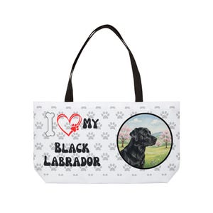 Może przedstawiać: Biała torba typu tote z czarnymi uchwytami. Zawiera ilustrację czarnego labradora w okrągłej ramce. Torba ma napis "I love my Black Labrador" z odciskami łap i kością.
