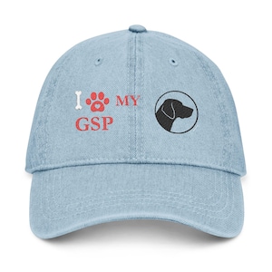 May include: Light blue denim baseball cap with embroidered text and graphics. The text reads "I ❤️ MY GSP" in white and red. A black silhouette of a dog's head is on the right side of the cap. The cap has a curved brim and an adjustable strap.