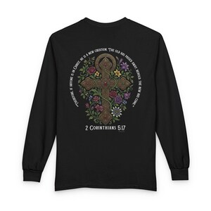 以下が含まれることがあります： 詳細な十字架のデザイン、花のアクセント、そして「2コリント5:17」のテキストが特徴の黒い長袖シャツ。十字架は円形のテキストで囲まれています。スタイリッシュで快適なアパレルアイテム。