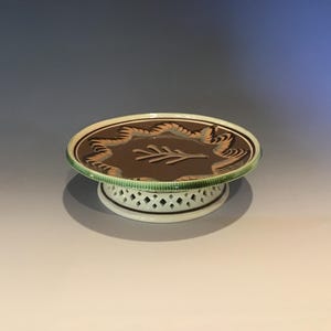 May include: A decorative ceramic cake stand with a brown top featuring a leaf design and a decorative border. The rim is green, and the base is white with a lattice pattern. The stand is elevated.