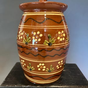 May include: A brown ceramic jar with a lid, decorated with horizontal bands, wavy lines, and floral patterns. The jar has a rounded body and a small knob on the lid. The design includes white, black, and green accents. This decorative jar is suitable for kitchen storage.