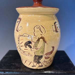 May include: A tan ceramic jar with a lid, featuring a brown knob. The jar is decorated with a painted scene of a person and a dog. The jar is sitting on a black surface.