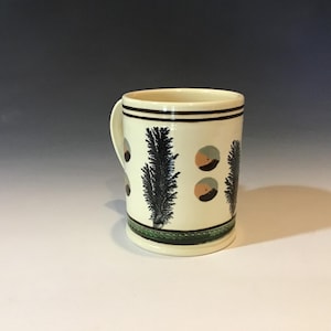 May include: A ceramic mug with a cream-colored base, featuring black and blue fern-like designs and small, circular accents in shades of orange and gray. The mug has a dark brown rim and handle, and a green band at the base.